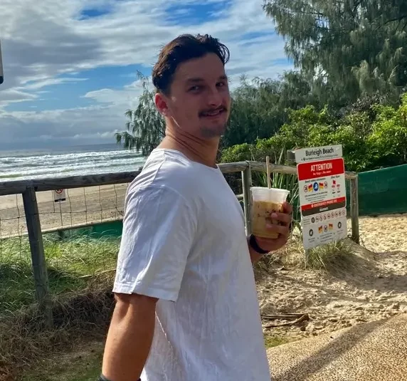 A man in a white shirt and denim shorts walks along a beach path, holding a drink. He has a wrist brace on his left hand. In the background, there's a sign for a beach vehicle access ramp and lush greenery. The sky is partly cloudy.